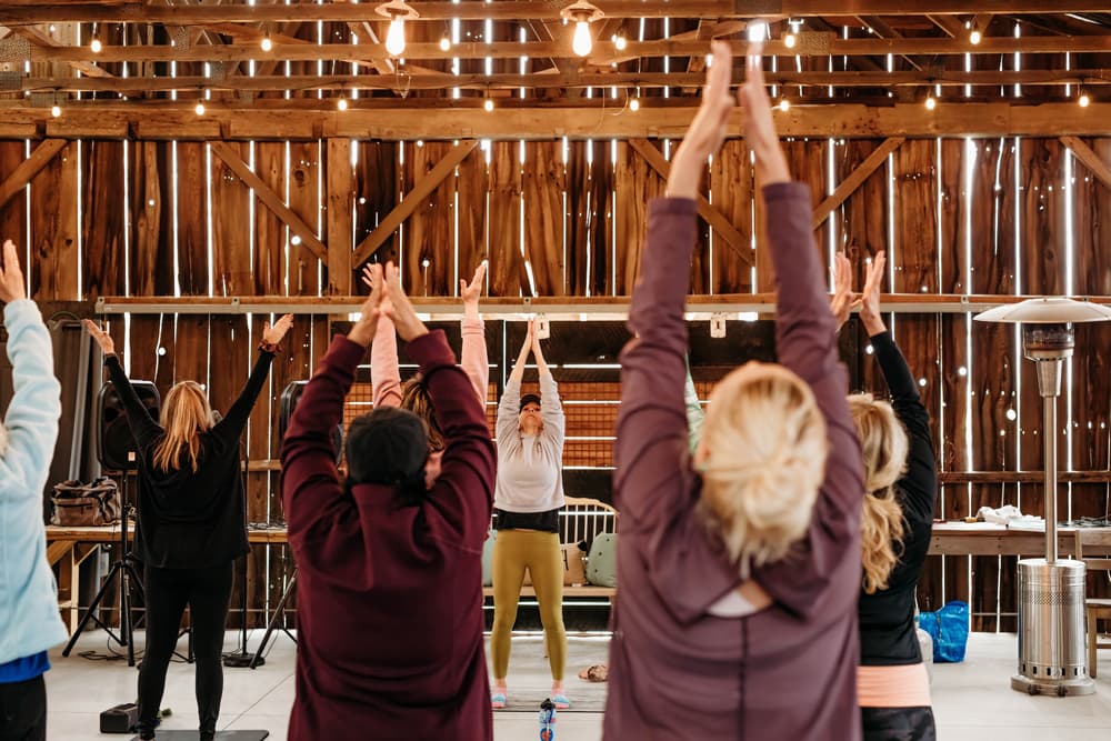 Seven women, all wearing black tops, smile while holding a plank position in a bright, spacious fitness studio with wooden floors, brick walls, and dumbbells stacked on shelves in the background.